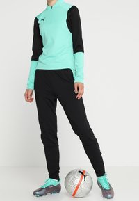 Mintgrön träningsskjorta med svarta ärmar, dragkedjekrage; svarta byxor; silvriga och teal fotbollsskor; strukturerad fotboll i närheten.