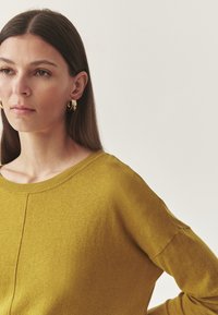 Sárga kötött pulóver széles nyakkivágással és ejtett vállú ujjal. A modellen arany karika fülbevaló van, haját egyenesen viseli, amely az egyik vállára omlik.