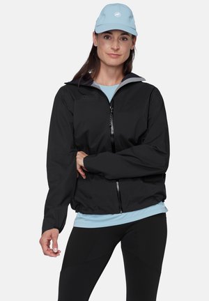 Vrouw draagt een zwart jack met rits, lichtblauw petje en shirt, en zwarte broek, staat met één arm over haar lichaam tegen een effen achtergrond.