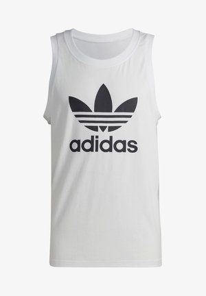 Mann trägt weißes ärmelloses Adidas-Shirt, schwarze Adidas-Shorts mit Streifen, gestreifte schwarze Socken und weiße Turnschuhe, steht mit den Händen in den Taschen.