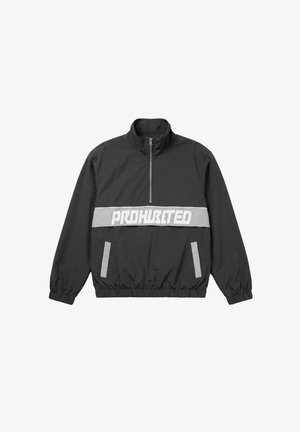 Junger Mann trägt eine dunkelgrüne Mütze, eine schwarze Jacke mit dem Text "PROHWRLTD" und ein kariertes Hemd, posiert vor weißem Hintergrund.