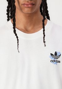 Hvit bomulls t-skjorte med en svart Adidas-logo med blå blomsteraksenter på venstre bryst. Rund hals med en jevn tekstur.