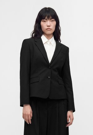 Femme aux longs cheveux noirs portant un blazer noir sur une chemise blanche à col, devant un fond clair uni.