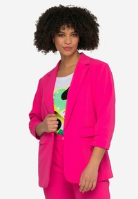Blazer rose vif au design ajusté, avec un col à revers et des manches retroussées, fabriqué à partir d'un tissu lisse. Porté par-dessus un t-shirt graphique coloré.