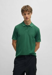 Grön polo-shirt med krage, korta ärmar och en diskret logotyp på bröstet. Tyget ser slätt och texturerat ut.