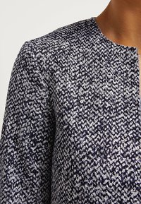 Detailní záběr osoby, která má na sobě texturovanou pletenou bundu navy a bílé barvy s kulatým výstřihem, kde jsou vidět ramena a horní část hrudn�íku.