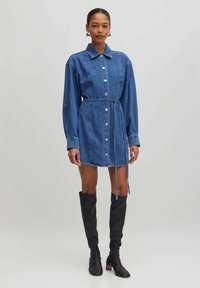Denim shirtjurk met lange mouwen, knoopsluiting en verstelbare tailleband. Kenmerken een lichte blauwe wassing en een rechte zoom. Gecombineerd met kniehoge zwarte laarzen.