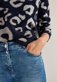 Marineblauer, flauschiger Pullover mit einem cremefarbenen Leopardenmuster, kombiniert mit hellblauen, verwaschenen Jeans, die einen Doppelknopfverschluss und eine Tasche aufweisen.