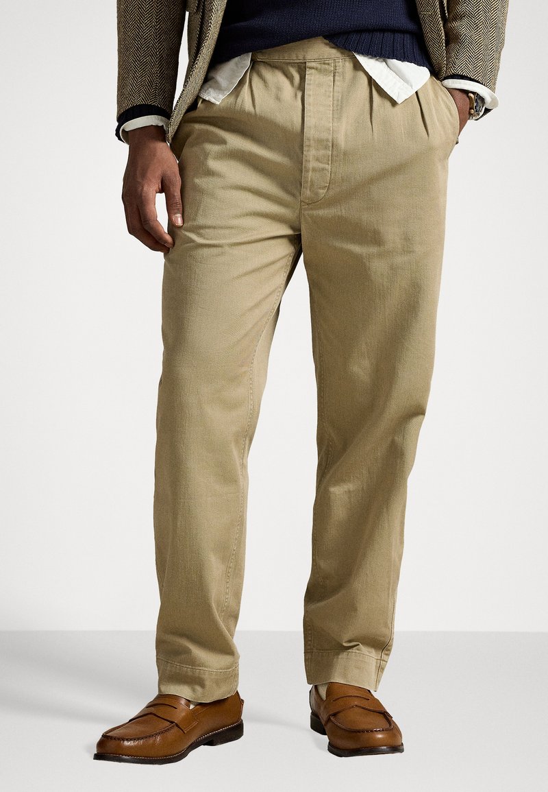 Pantalons plissés kaki à coupe décontractée, fabriqués en tissu tissé. Associés à des mocassins marron, affichés sur un fond uni.