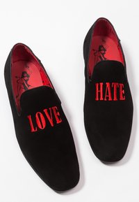 Een paar zwarte suède instapschoenen met rode borduursels met de tekst "LOVE" aan de linkerkant en "HATE" aan de rechterkant, met rode zolen met een afbeelding van een duivelse vrouw.