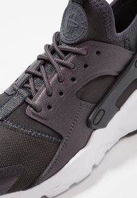 Mustat ja harmaat urheilukengät, joissa on tekstiili- ja synteettisiä materiaaleja, ulkonäöltään teksturoitu yläosa, nauhakiinnitys ja valkoinen kumipohja.