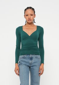 Grünes Langarmoberteil mit tiefem V-Ausschnitt, geraffter Textur an der Taille und taillierter Silhouette, kombiniert mit hellblauen hochgeschnittenen Jeans.