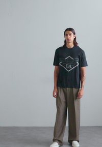 Svart t-skjorte med kort erme og geometrisk logo, kombinert med løse olivengrønne bukser og hvite joggesko. Enkelt design, avslappet passform.