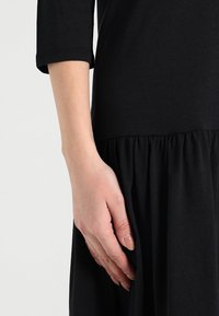 Robe noire avec un col arrondi, des manches trois-quarts et une jupe froncée. Gros plan sur une main posée sur le tissu.