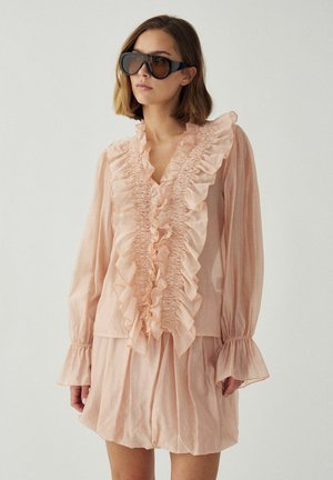 Žena v svetloružovej blúzke s volánmi a dlhými rukávmi, v rovnakých sukňových nohaviciach, doplnená veľkými tmavými slnečnými okuliarmi.