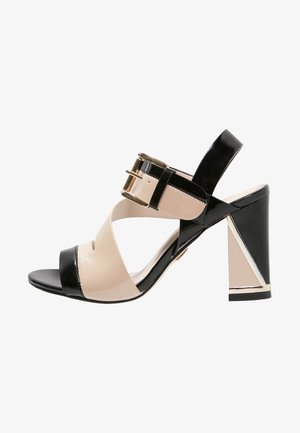 Sandalia de tacón alto con punta abierta, con tiras de charol beige y negro, y una hebilla grande dorada en la correa del tobillo.