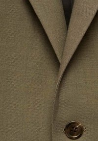 Olivgrüner Blazerstoff mit strukturiertem Finish, ausgestattet mit einem großen Knopfdetail und spitzen Revers. Nahaufnahme des Kleidungsstücks.