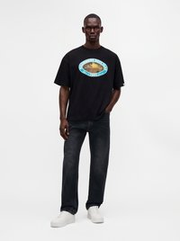 Man wearing black Billionaire Boys Club t-shirt, dark jeans, and white sneakers standing with one hand in pocket against white background.