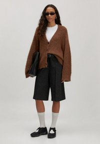 Braune, flauschige Strickjacke mit Knöpfen, weißes T-Shirt, dunkle Shorts, schwarze Sneakers und weiße Socken. Eine schwarze Tasche und Sonnenbrille runden den Look ab.