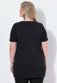 Mujer con cabello largo y rubio, vistiendo una camiseta negra de manga corta lisa y pantalones negros, de pie frente a una pared blanca.