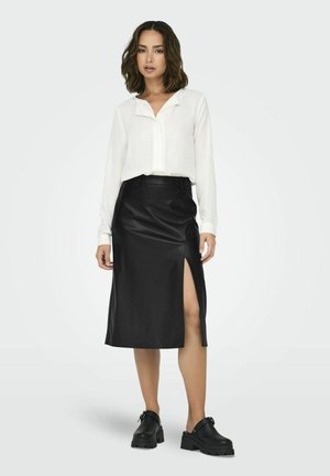 Jupe midi en cuir noir avec une fente latérale, associée à une chemise blanche à boutons. La jupe présente une texture lisse et une coupe ajustée.
