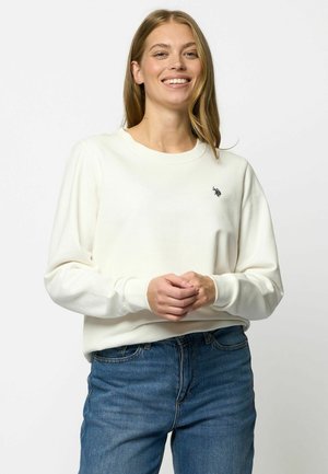 Hvit langermet genser med rund hals og ribbestrikkede mansjetter, prydet med en liten brodert logo på brystet, sammen med blå denimjeans.