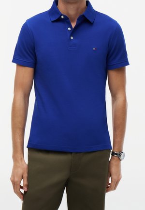 Homme portant un polo bleu roi à manches courtes avec des boutons, assorti à un pantalon vert olive et une montre-bracelet argentée au poignet gauche.