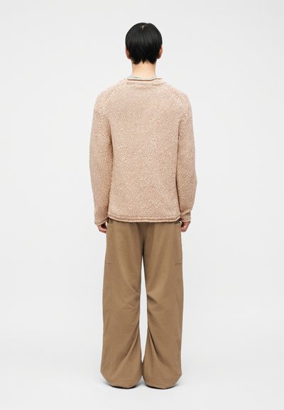 Personne debout de dos, portant un pull beige texturé et un pantalon marron ample, sur un fond clair uni.