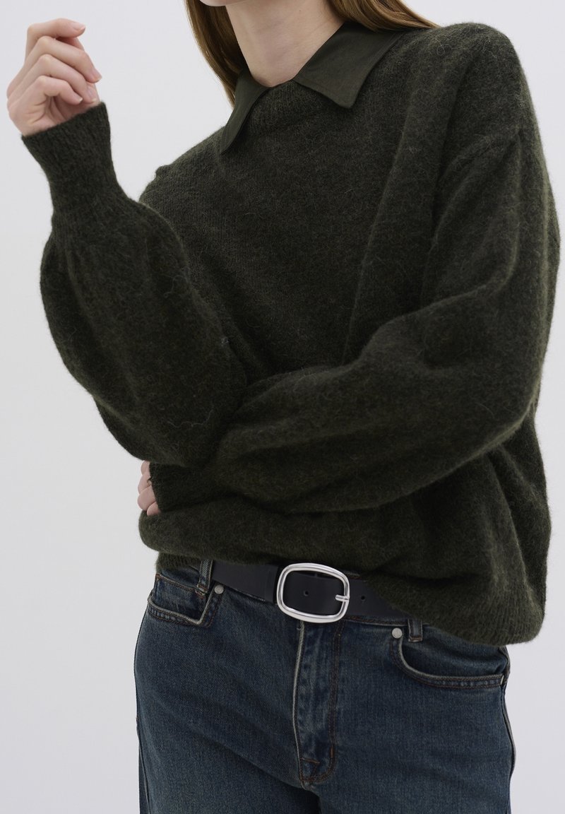 Femme portant un pull duveteux vert foncé à manches ballon, une chemise noire à col en dessous, un jean bleu et une ceinture noire avec une boucle argentée.