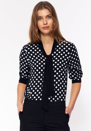 Czarna i biała bluzka w kropki z wiązanym dekoltem, rękawami o długości trzech czwartek oraz dopasowanym krojem. Gładki materiał z kontrastowymi akcentami.