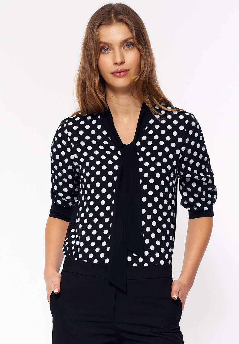 Czarna i biała bluzka w kropki z wiązanym dekoltem, rękawami o długości trzech czwartek oraz dopasowanym krojem. Gładki materiał z kontrastowymi akcentami.