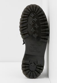 Semelle de botte noire rugueuse avec des rainures profondes et des motifs en forme de losange, inclinée contre une surface blanche d'angle.