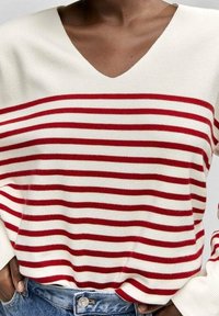 Henkilö, joka on pukeutunut valkoiseen V-päähineeseen, jossa on vaakasuoria punaisia raitoja, ja se on tungettu sinisiin farkkuihin, kädet taskuissa.