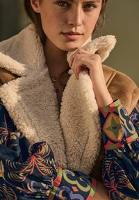 Tan shearling-takki, jossa on teksturoitu vaalean beige kaulus, yhdistettynä värikkääseen kuvioituun paitaan, jossa on sinisiä, vihreitä ja punaisia yksityiskohtia.