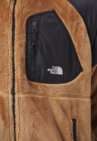 La giacca in pile presenta un tessuto marrone testurizzato, una tasca sul petto in nylon nero con cerniera e un logo bianco. La cerniera frontale è centrale.