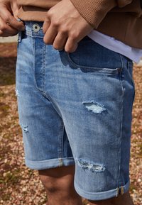 Shorts de mezclilla en azul claro con detalles de desgaste, dobladillo enrollado, bolsillos delanteros y cierre de botón metálico. Tejido texturizado con marcas de uso visibles.