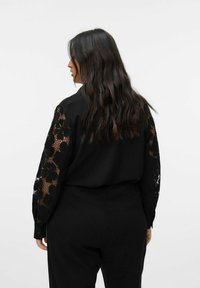 Blusa nera con maniche lunghe e dettagli in pizzo floreale, chiusura frontale con bottoni e vestibilità comoda. Il tessuto sembra liscio e leggero.