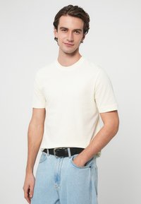 Kremowy bawełniany t-shirt z krótkimi rękawami, okrągłym dekoltem, noszony z jasnoniebieskimi dżinsami i czarnym paskiem. Gładka faktura, casualowy krój.