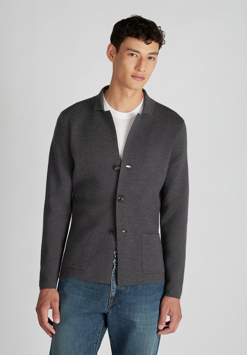 Grau gestrickte Strickjacke mit Schalkragen, fünf Knöpfen und zwei vorderen Taschen. Über einem weißen T-Shirt getragen und mit blauen Jeans kombiniert.