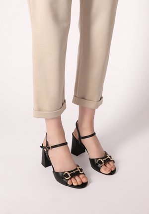 Schwarze Leder-Sandalen mit Blockabsatz verfügen über eine offene Zehenpartie und einen Knöchelriemen mit goldenen Schnallenakzenten. Die Sohle ist glatt und elegant.