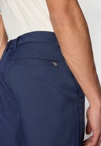 Σκούρα μπλε παντελόνια από λείο ύφασμα, με πίσω τσέπη που διαθέτει μικρή ετικέτα λογότυπου. Φτιαγμένα σε εφαρμοστή γραμμή με θηλιές για ζώνη και κομψό σχεδιασμό.