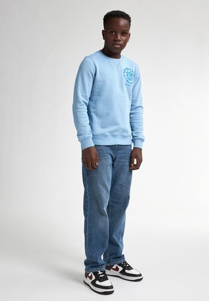 Sweatshirt bleu clair avec un col rond et un logo sur le côté gauche de la poitrine, associé à un jean droit bleu et des baskets noir et blanc.