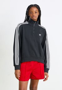 Juodas zip up džemperis su baltais trimis dryžiais ant rankovių, turintis juostuotą apačią ir mažą baltą logotipą ant krūtinės. Derinamas su raudonomis šortais.
