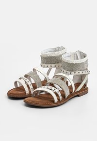White flat sandals with multiple textured straps, silver studs, and rhinestone embellishments on brown soles.