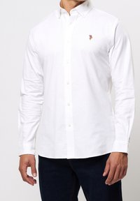 Biała koszula z długim rękawem, kołnierzykiem na guziki, patką na przodzie oraz haftowanym akcentem na piersi. Materiał z mieszanki bawełny, gładka faktura.