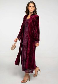 Longue veste en velours bordeaux au-dessus d'une robe légère violette foncée à motifs floraux, associée à des sandales à talons aiguilles nudes et à une pochette.