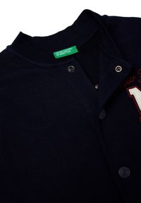 Námořnicky modrý kardigan s žebrovaným límcem a zapínáním na patentky. Má vyšívaný detail "New York" a zelenou značkovou štítek. Měkká, strukturovaná látka.