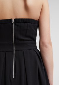 Zwarte strapless jurk met een aangesloten lijfje, een geplooide rok en een zichtbare rugrits met een zilveren trekker. Gladde stoftextuur.