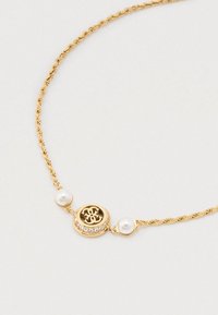 Złoty splątany naszyjnik ma centralny okrągły wisiorek z czarnym wzorem, otoczony przez przezroczyste kamienie i dwie białe perły.