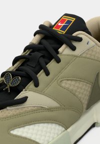 Nike kedas ar brūnu, zaļu un melnu krāsu shēmu. Izceļas ar teksturētām materiālu virsmām, kontrastējošiem auklām un zelta tenisa raketes akcentu.